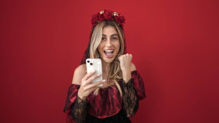 Foto de Mujer rubia joven usando teléfono inteligente que tiene fiesta de Halloween sobre fondo rojo aislado - Imagen libre de derechos