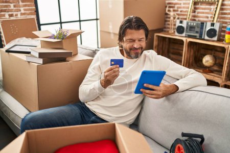 Photo for Middle age man using touchpad and credit card sitting on sofa at new home - Royalty Free Image