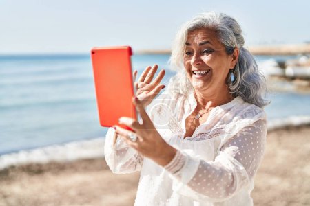 Foto de Mujer de mediana edad sonriendo confiado teniendo videollamada en la playa - Imagen libre de derechos