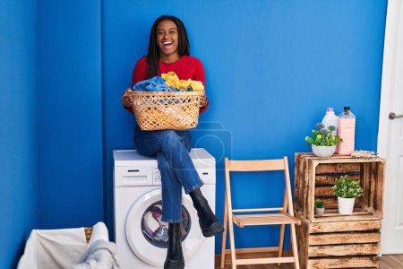 Téléchargez les photos : Jeune afro-américain avec des tresses tenant panier à linge assis sur la machine à laver souriant et riant fort à haute voix parce que drôle blague folle. - en image libre de droit