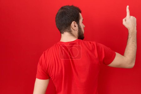 Foto de Joven hombre hispano con camiseta roja casual posando hacia atrás apuntando hacia adelante con la mano del dedo - Imagen libre de derechos