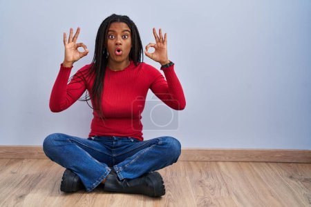 Photo for Young african american with braids sitting on the floor at home looking surprised and shocked doing ok approval symbol with fingers. crazy expression - Royalty Free Image