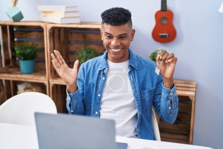 Photo for Young hispanic man holding keys of new home doing video call celebrating achievement with happy smile and winner expression with raised hand - Royalty Free Image