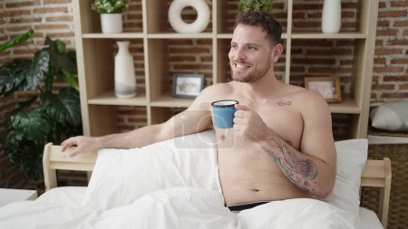 Foto de Joven hombre caucásico bebiendo un café sentado en la cama sin camisa en el dormitorio - Imagen libre de derechos