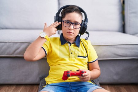 Foto de Joven niño hispano jugando videojuego sosteniendo controlador usando auriculares disparando y matándose apuntando de la mano y los dedos a la cabeza como arma, gesto suicida. - Imagen libre de derechos