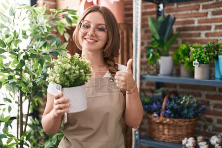 Foto de Mujer joven caucásica trabajando en floristería sosteniendo planta sonriendo feliz y positivo, pulgar hacia arriba haciendo excelente y signo de aprobación - Imagen libre de derechos