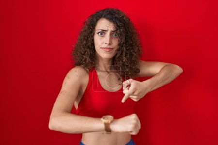 Téléchargez les photos : Femme hispanique avec les cheveux bouclés debout sur fond rouge à la hâte pointant vers regarder le temps, impatience, bouleversé et en colère pour retard de délai - en image libre de droit