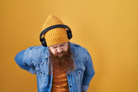 Photo for Caucasian man with long beard listening to music using headphones suffering of backache, touching back with hand, muscular pain - Royalty Free Image