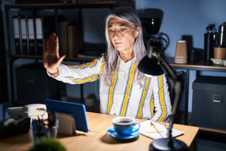 Téléchargez les photos : Femme d'âge moyen avec les cheveux gris travaillant au bureau la nuit faire arrêter de chanter avec la paume de la main. expression d'avertissement avec geste négatif et sérieux sur le visage. - en image libre de droit