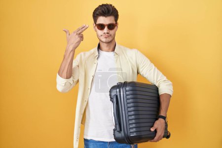 Foto de Joven hispano sosteniendo maleta yendo de vacaciones de verano disparando y matándose apuntando de la mano y los dedos a la cabeza como arma, gesto suicida. - Imagen libre de derechos