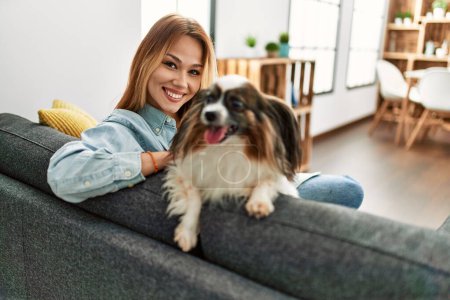 Téléchargez les photos : Jeune femme caucasienne souriante confiante assise sur un canapé avec chien à la maison - en image libre de droit