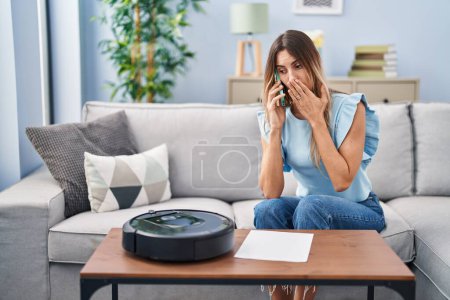 Téléchargez les photos : Jeune femme hispanique assise à la maison par aspirateur robot parlant au téléphone couvrant la bouche avec la main, choquée et effrayée par l'erreur. expression surprise - en image libre de droit