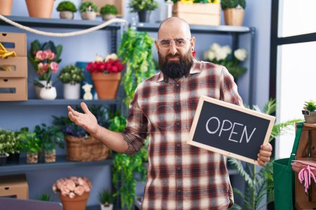 Photo for Young hispanic man with beard and tattoos working at florist holding open sign clueless and confused expression. doubt concept. - Royalty Free Image