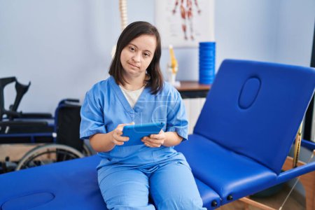 Foto de Síndrome de Down mujer que usa uniforme de fisioterapia usando touchpad en la clínica de fisioterapia - Imagen libre de derechos