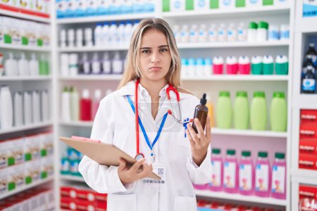 Téléchargez les photos : Jeune femme blonde travaillant à la pharmacie pharmacie tenant bouteille sceptique et nerveux, fronçant les sourcils bouleversés en raison de problèmes. personne négative. - en image libre de droit