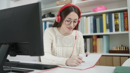 Téléchargez les photos : Jeune belle étudiante hispanique utilisant ordinateur et écouteurs écrit sur ordinateur portable à l'université de la bibliothèque - en image libre de droit