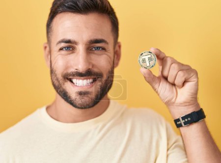 Schöner hispanischer Mann, der eine Kryptowährungsmünze in der Hand hält, sieht positiv und glücklich stehend aus und lächelt mit einem selbstbewussten Lächeln, das Zähne zeigt 