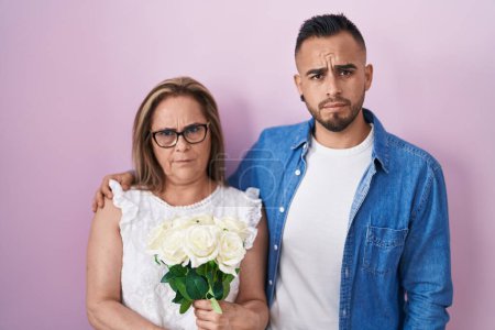 Téléchargez les photos : Mère hispanique et fils tenant ensemble bouquet de fleurs blanches sceptiques et nerveux, fronçant les sourcils bouleversés en raison de problèmes. personne négative. - en image libre de droit