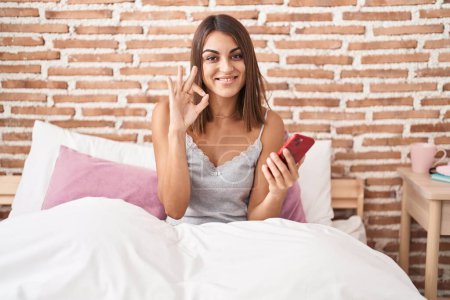 Téléchargez les photos : Jeune femme hispanique à l'aide d'un smartphone assis sur le lit à la maison faisant signe ok avec les doigts, sourire geste amical excellent symbole - en image libre de droit