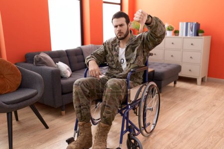 Téléchargez les photos : Jeune soldat hispanique blessé assis sur un fauteuil roulant avec un visage en colère, signe négatif montrant aversion avec les pouces baissés, concept de rejet - en image libre de droit