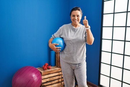 Photo for Middle age hispanic woman holding pilates ball smiling happy and positive, thumb up doing excellent and approval sign - Royalty Free Image