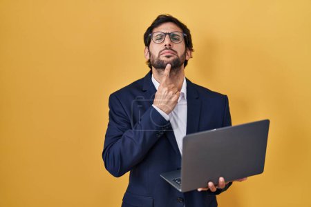 Foto de Hombre latino guapo trabajando usando computadora portátil pensando concentrado en la duda con el dedo en la barbilla y mirando hacia arriba preguntándose - Imagen libre de derechos