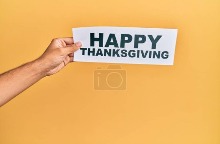 Hand of caucasian man holding paper with happy thanksgiving message over isolated yellow background