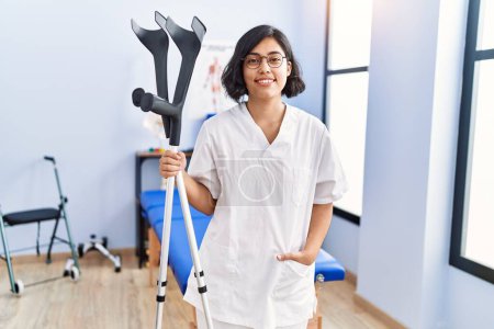 Téléchargez les photos : Jeune femme latine portant un uniforme de physiothérapeute tenant des béquilles à la clinique de physiothérapie - en image libre de droit