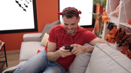 Photo for Young caucasian man wearing devil costume sitting on the sofa using smartphone at home - Royalty Free Image