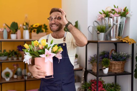 Foto de Hombre de mediana edad con barba trabajando en floristería sosteniendo la planta sonriendo feliz haciendo signo de bien con la mano en el ojo mirando a través de los dedos - Imagen libre de derechos