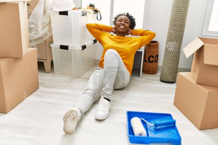 Téléchargez les photos : Femme afro-américaine assise sur le sol détendue avec les mains sur la tête à la nouvelle maison - en image libre de droit