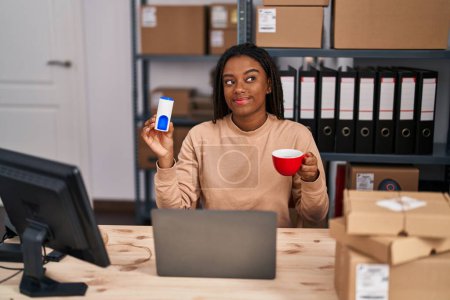 Téléchargez les photos : Jeune afro-américain avec des tresses travaillant dans une petite entreprise e-commerce boire un café avec édulcorant souriant regardant vers le côté et regardant loin en pensant. - en image libre de droit