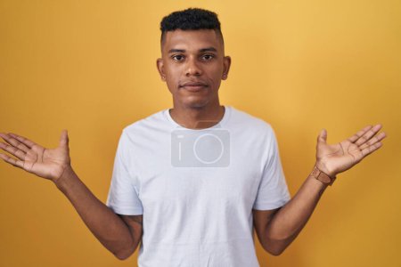 Foto de Young hispanic man standing over yellow background clueless and confused expression with arms and hands raised. doubt concept. - Imagen libre de derechos