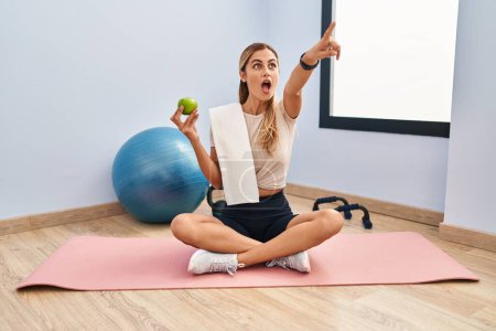 Photo for Young blonde woman wearing sportswear and towel holding healthy apple pointing with finger surprised ahead, open mouth amazed expression, something on the front - Royalty Free Image