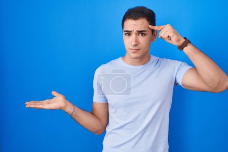 Photo for Young hispanic man standing over blue background confused and annoyed with open palm showing copy space and pointing finger to forehead. think about it. - Royalty Free Image
