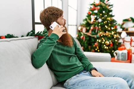 Foto de Young redhead man talking on the smartphone sitting by christmas tree at home - Imagen libre de derechos