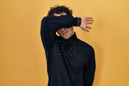 Téléchargez les photos : Homme hispanique debout sur fond jaune couvrant les yeux avec le bras, l'air grave et triste. concept de non-visibilité, de dissimulation et de rejet - en image libre de droit