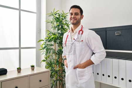 Foto de Joven hombre hispano vistiendo uniforme médico sonriendo confiado en la clínica - Imagen libre de derechos