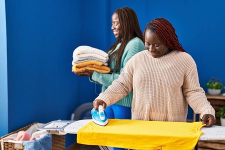 Foto de Mujeres afroamericanas planchando camiseta sosteniendo ropa doblada en la lavandería - Imagen libre de derechos