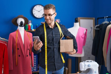 Téléchargez les photos : Jeune homme caucasien tailleur en utilisant smartphone tenant paquet de livraison à l'usine de vêtements - en image libre de droit