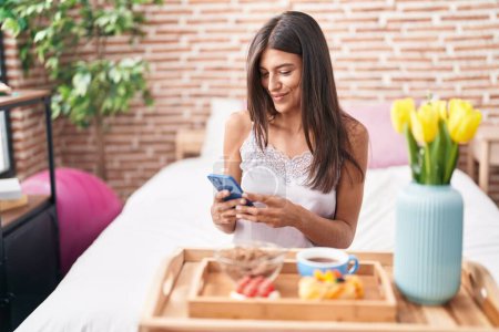 Photo for Young beautiful hispanic woman having gift breakfast using smartphone at bedroom - Royalty Free Image