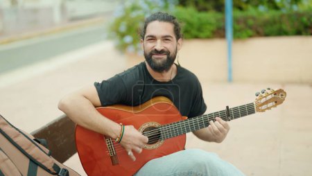 Foto de Joven músico hispano tocando la guitarra clásica sentado en el banco del parque - Imagen libre de derechos