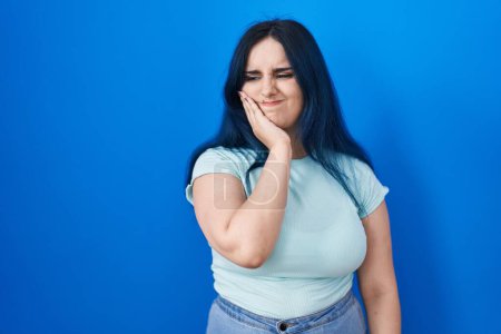 Photo for Young modern girl with blue hair standing over blue background touching mouth with hand with painful expression because of toothache or dental illness on teeth. dentist - Royalty Free Image
