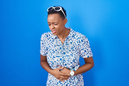 Téléchargez les photos : African american woman standing over blue background with hand on stomach because indigestion, painful illness feeling unwell. ache concept. - en image libre de droit