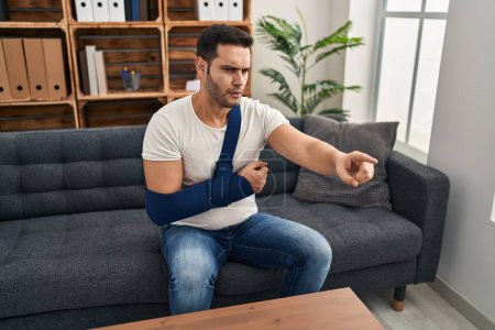 Photo for Young hispanic man with beard wearing arm on sling sitting at therapy consult pointing with finger to the camera and to you, confident gesture looking serious - Royalty Free Image