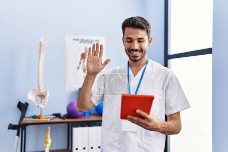 Photo for Young hispanic physiotherapist man doing online appointment looking positive and happy standing and smiling with a confident smile showing teeth - Royalty Free Image
