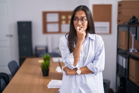 Foto de Mujer hispana joven en la oficina que parece estresada y nerviosa con las manos en la boca mordiéndose las uñas. problema de ansiedad. - Imagen libre de derechos