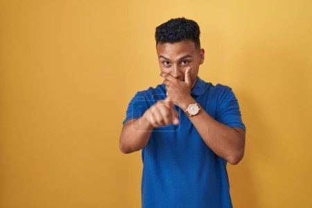 Téléchargez les photos : Jeune homme hispanique debout sur fond jaune se moquant de vous, pointant du doigt la caméra avec la main sur la bouche, expression de honte - en image libre de droit
