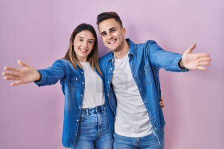 Téléchargez les photos : Jeune couple hispanique debout sur fond rose regardant la caméra souriant à bras ouverts pour un câlin. expression joyeuse embrassant le bonheur. - en image libre de droit