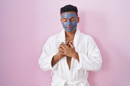 Téléchargez les photos : Jeune homme hispanique portant un masque de beauté et peignoir de bain souriant avec les mains sur la poitrine avec les yeux fermés et geste reconnaissant sur le visage. concept de santé. - en image libre de droit